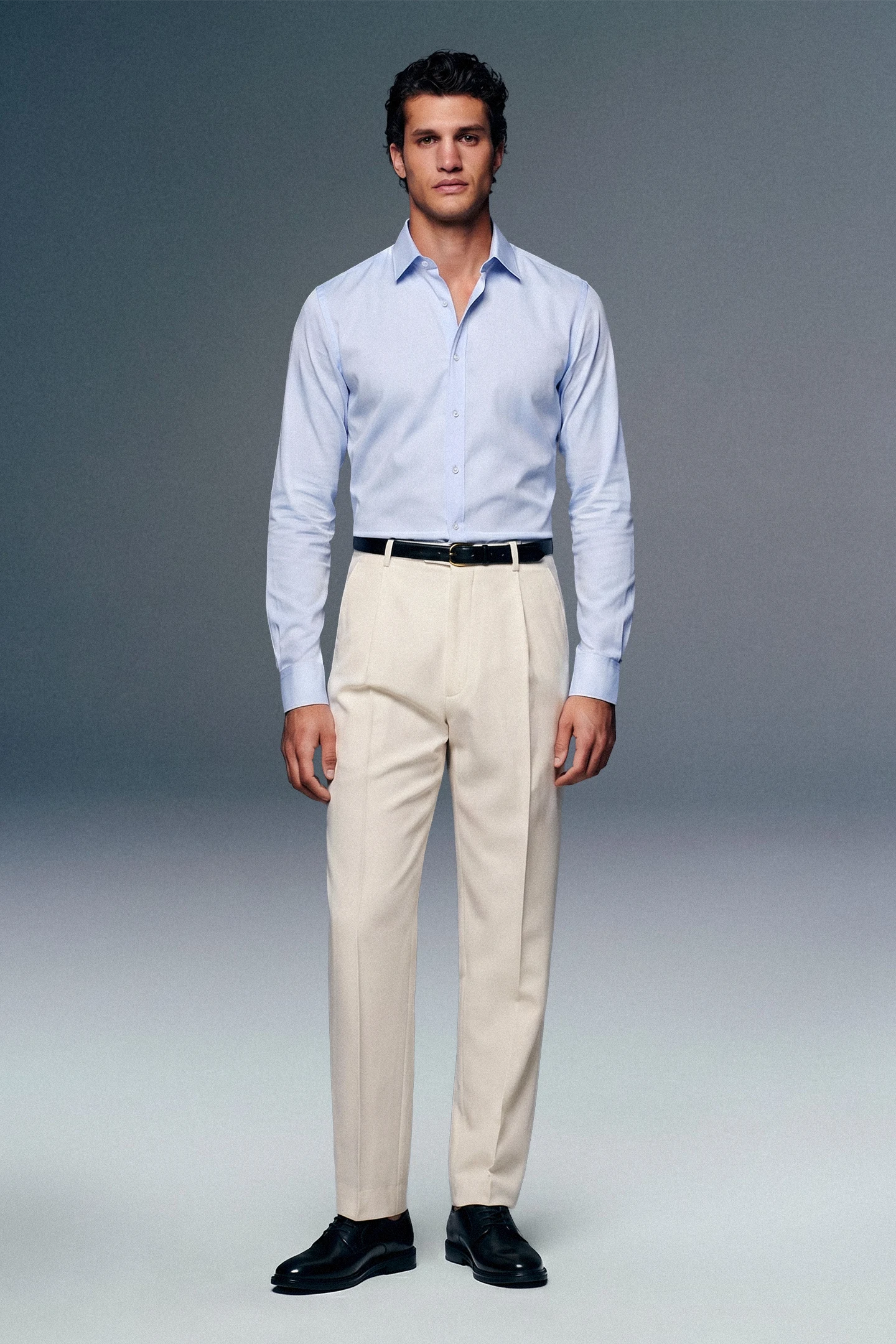 Man wearing a light blue dress shirt, beige pants, and black shoes on a gray background