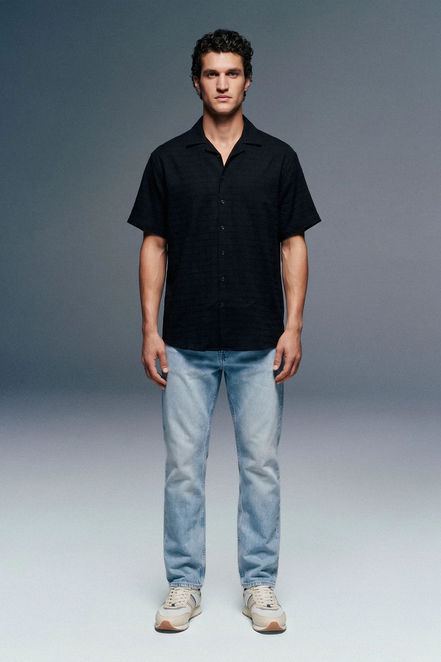Man wearing a black shirt and light blue jeans against a gray background