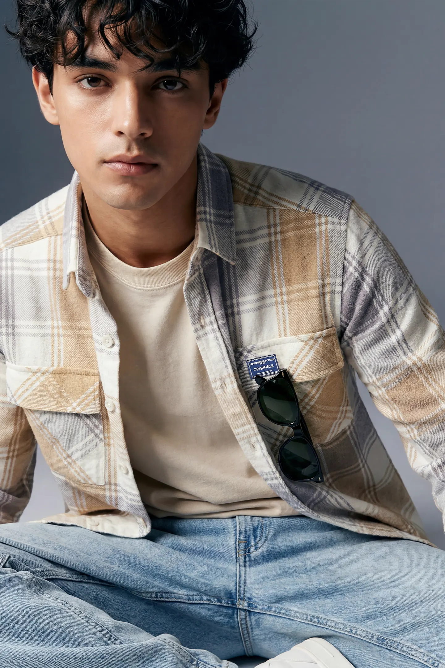 Man wearing a plaid shirt over a beige t-shirt with sunglasses in the pocket, sitting against a gray background.