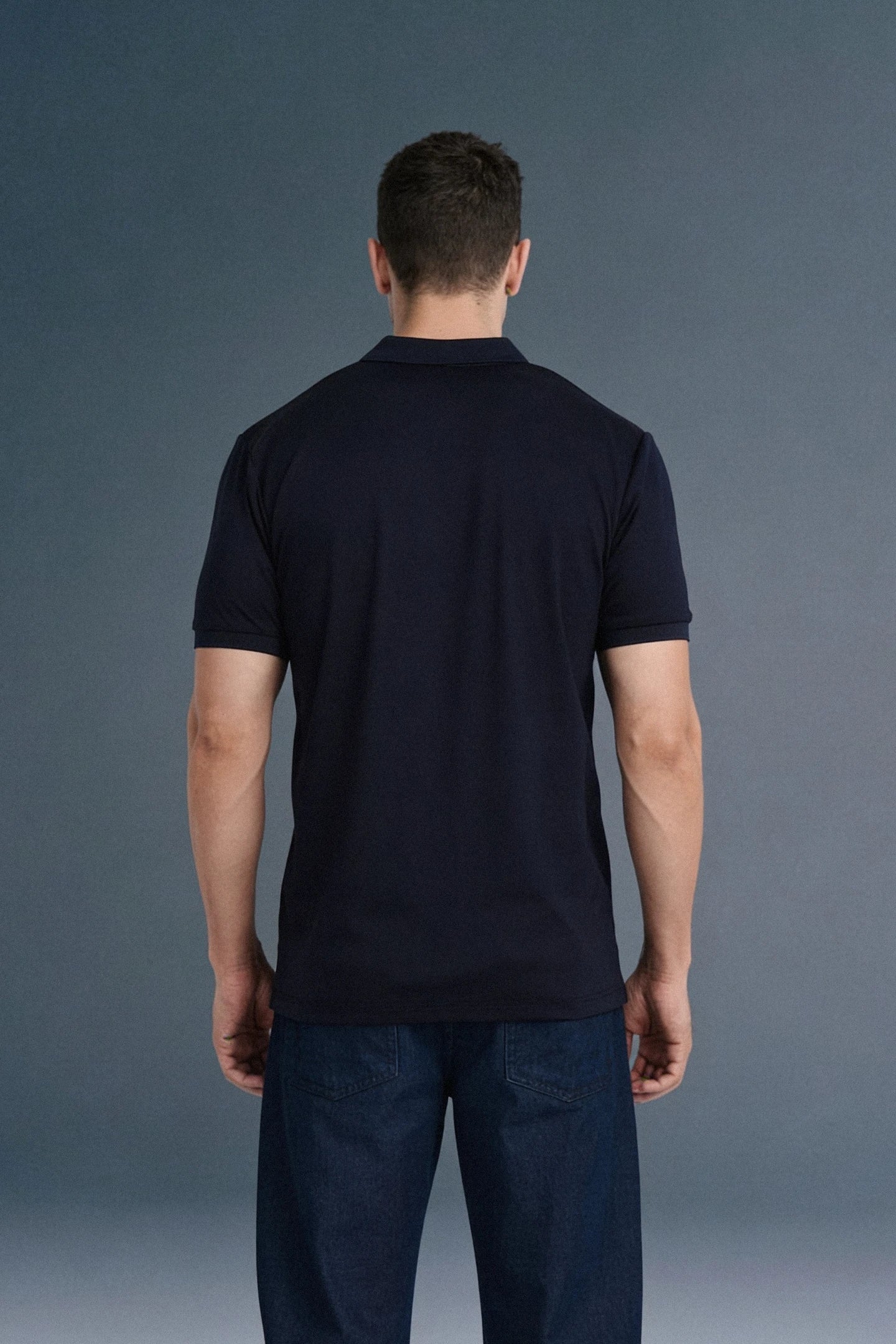 Full-length rear view of a man standing straight, showing the clean back fit of a solid navy blue polo shirt and dark blue trousers.