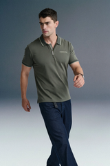 Full-length front view of a man walking forward at an angle, wearing an olive green polo shirt and dark blue jeans against a grey gradient background.
