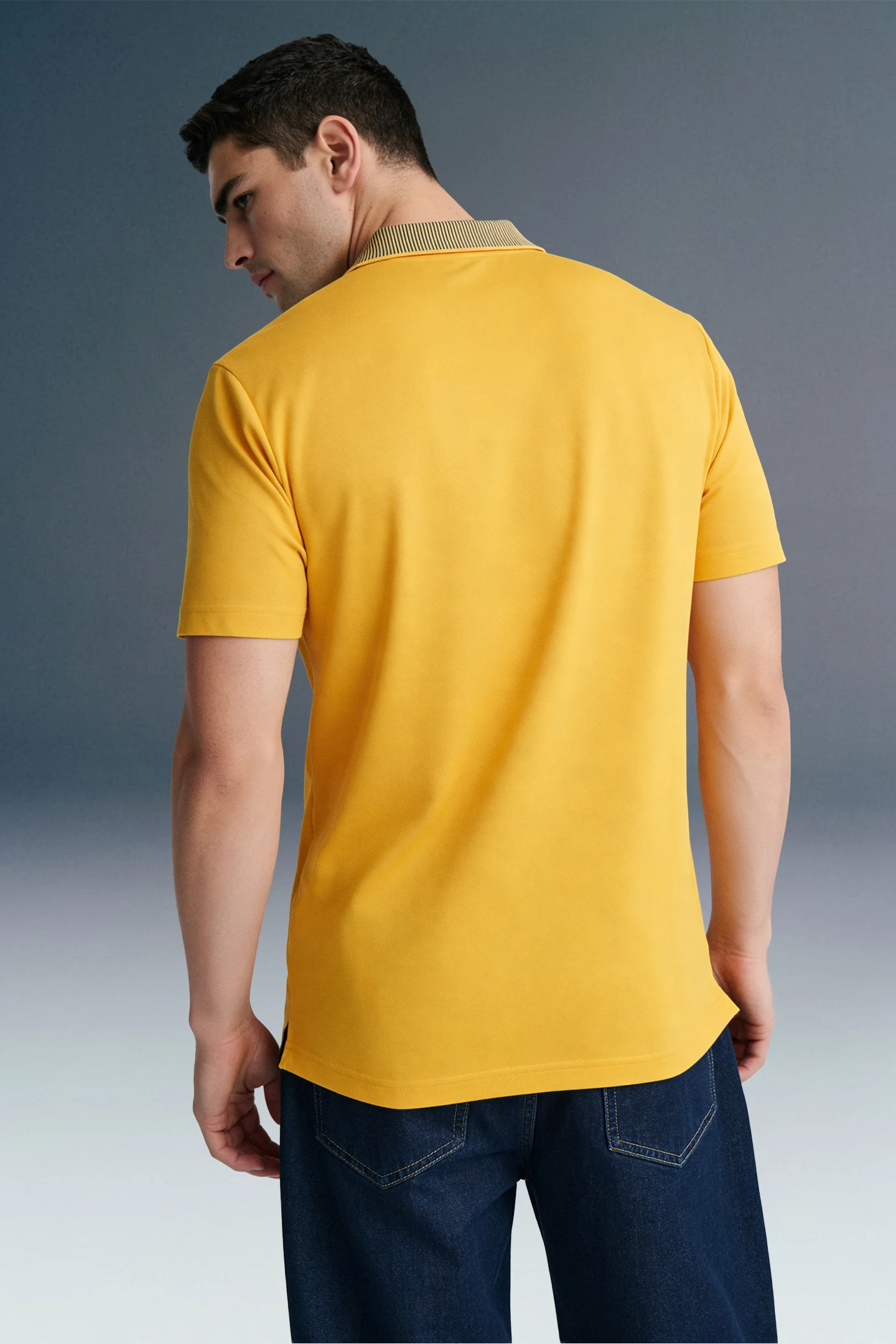 Rear view from the waist up of a man looking down and to his left, showcasing the fit of a mustard yellow polo shirt from behind.
