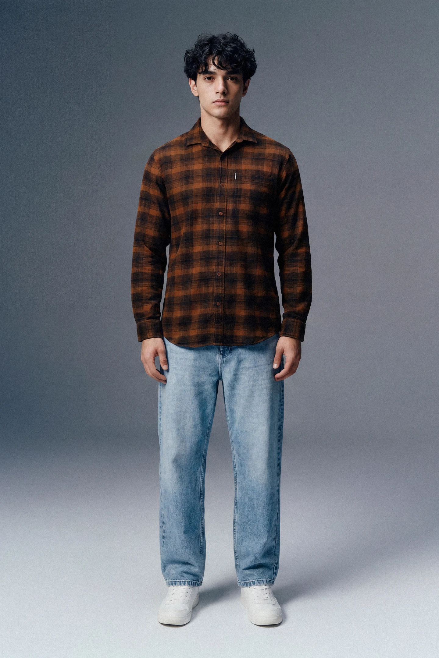 Man wearing a brown plaid shirt and light blue jeans against a gray background