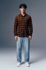 Man wearing a brown plaid shirt and light blue jeans against a gray background