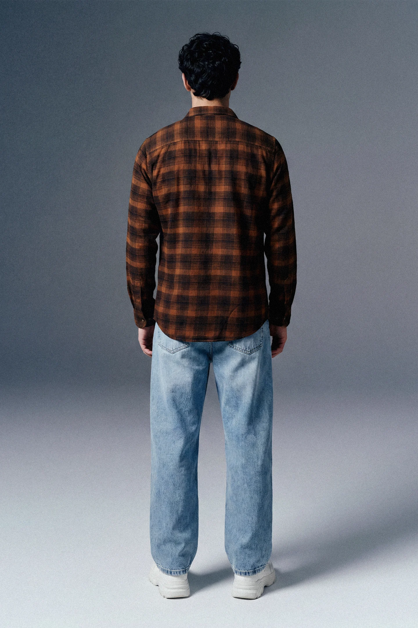 Person wearing a brown plaid shirt and light blue jeans against a gray background