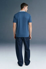 Full-length rear view of a man looking slightly down and to the right, wearing a slate blue polo shirt and dark trousers.