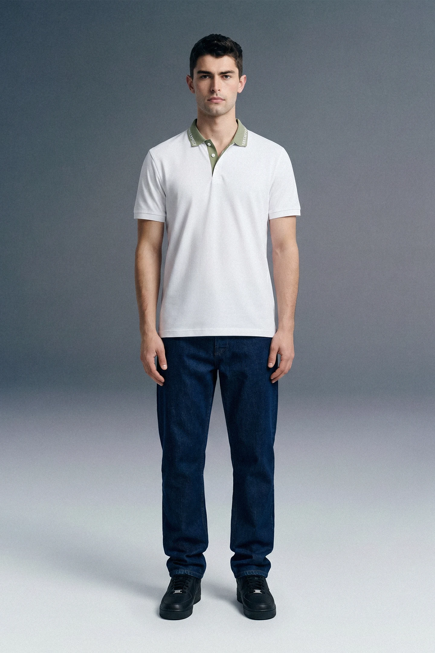 Full-length front view of a man standing straight and facing forward, wearing a white polo shirt with an olive green collar, paired with dark navy blue trousers.