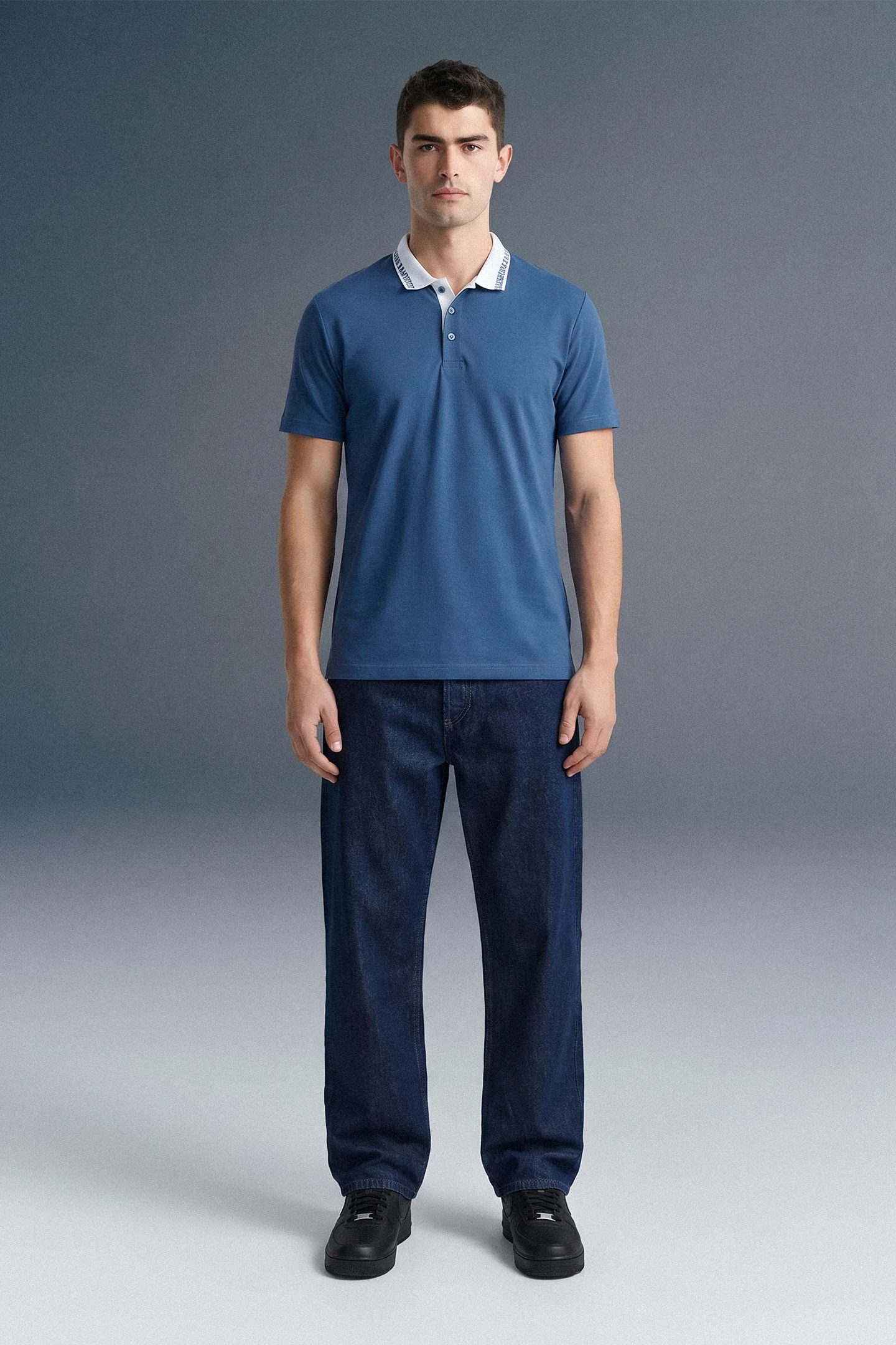Full-length front view of a man standing straight and facing forward, wearing a slate blue polo shirt with white collar detailing and dark navy blue pants.