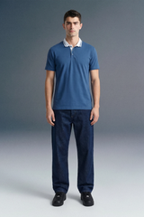 Full-length front view of a man standing straight and facing forward, wearing a slate blue polo shirt with white collar detailing and dark navy blue pants.