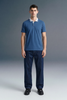 Full-length front view of a man standing straight and facing forward, wearing a slate blue polo shirt with white collar detailing and dark navy blue pants.
