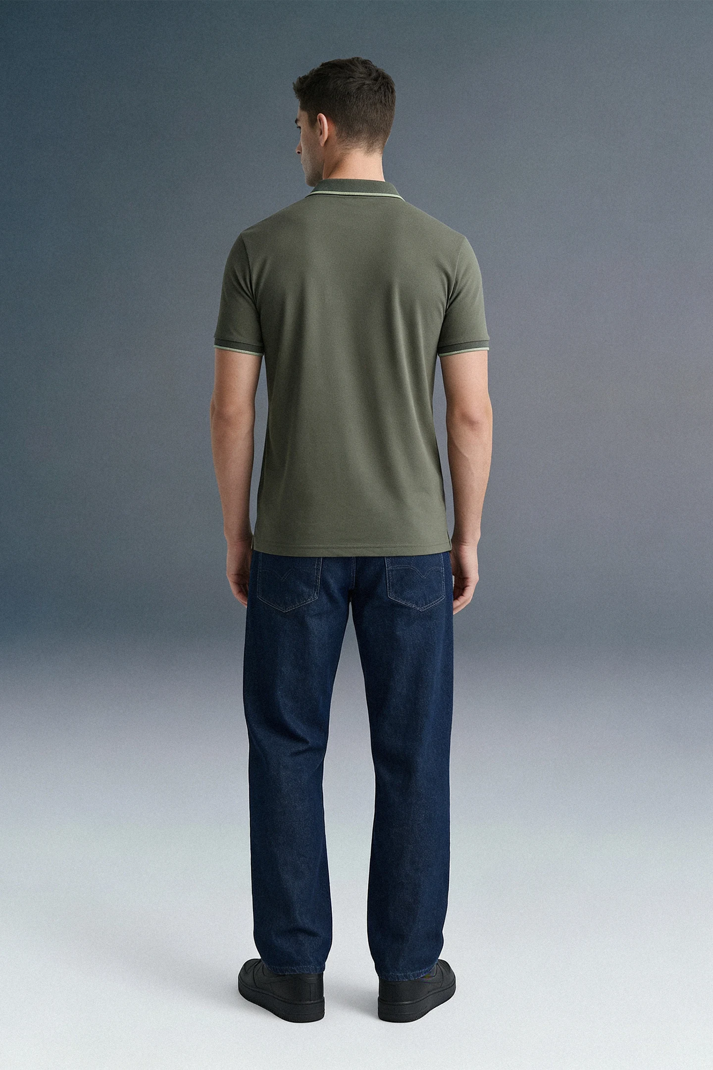 Full-length rear view of a man standing straight, wearing an olive green polo shirt featuring a thin light stripe on the collar, paired with dark navy blue trousers.