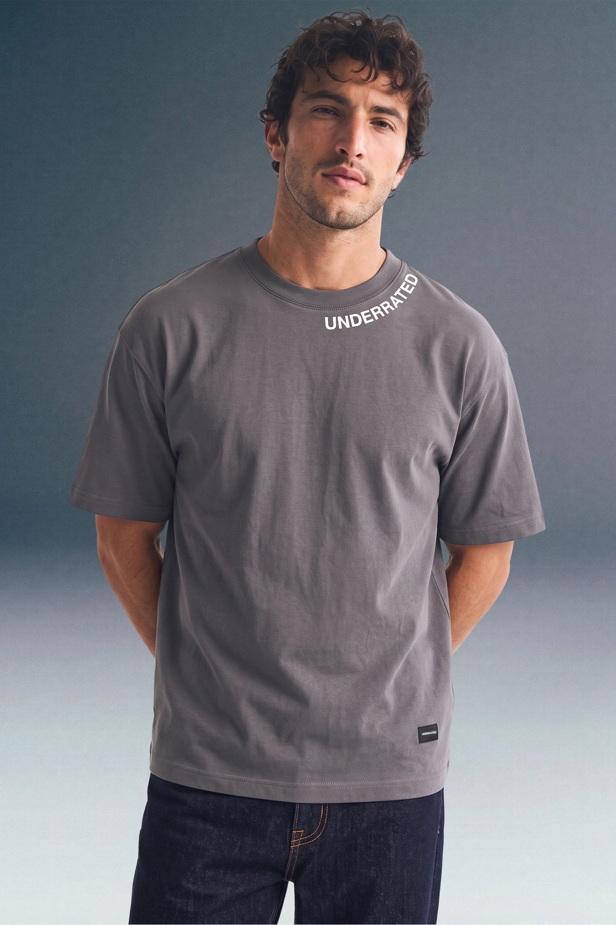 Front view from the waist up of a man standing with his hands behind his back, wearing a dark grey T-shirt with collar text detailing and a small brand patch near the left hem.