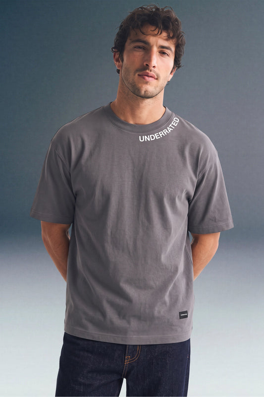 Front view from the waist up of a man standing with his hands behind his back, wearing a dark grey T-shirt with collar text detailing and a small brand patch near the left hem.