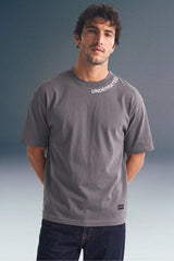 Front view from the waist up of a man standing with his hands behind his back, wearing a dark grey T-shirt with collar text detailing and a small brand patch near the left hem.