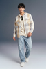 Man wearing a plaid shirt over a beige sweater with light blue jeans on a gray background