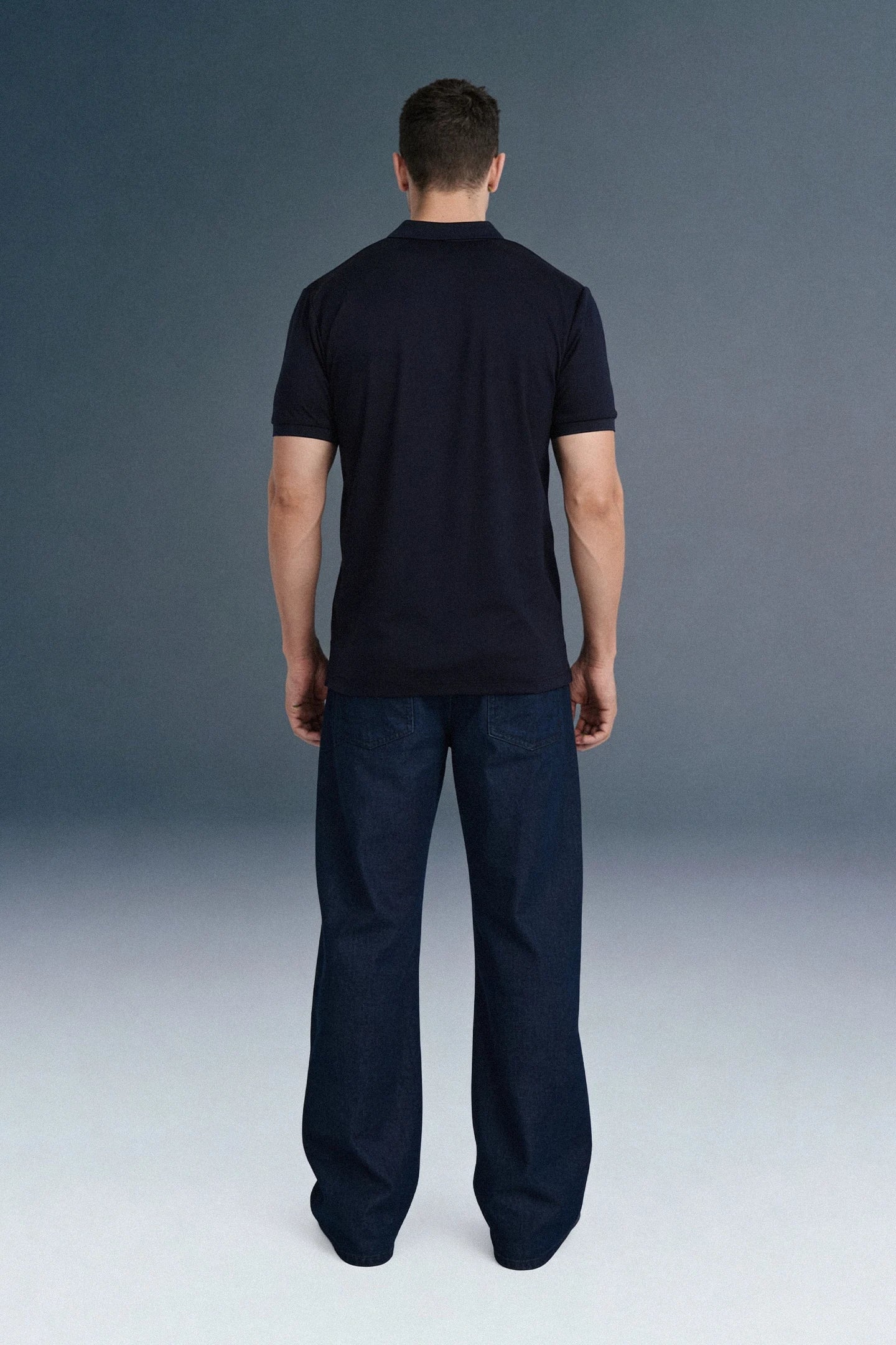 Full-length front view of a man mid-stride looking to his right, wearing a navy blue open-placket polo shirt and dark navy trousers.