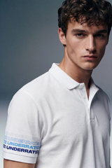 A close-up, front-facing portrait of a man looking directly ahead, showcasing the collar and the blue-patterned cuff detail on the sleeve of his white polo shirt.