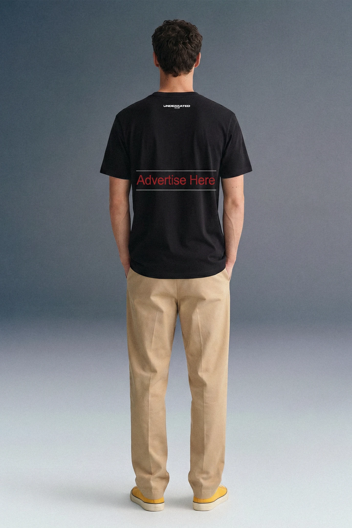 Full-length rear view of a man standing straight, showing the back of a black T-shirt featuring horizontal red branding text across the middle back, paired with khaki pants and yellow shoes.