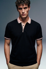 A medium close-up, front-facing shot of a man wearing a black polo shirt featuring a beige collar and patterned trims on the sleeve cuffs, paired with khaki trousers.