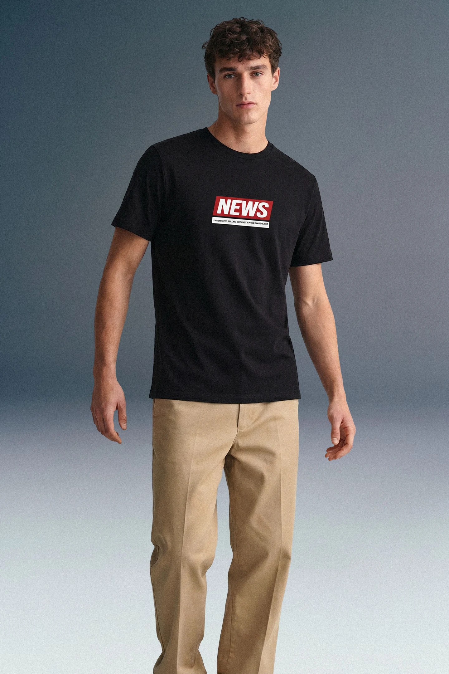 Full-length front view of a man standing against a grey studio backdrop, wearing a black T-shirt with a red and white rectangular graphic on the chest and beige khaki trousers.