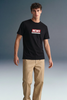 Full-length front view of a man standing against a grey studio backdrop, wearing a black T-shirt with a red and white rectangular graphic on the chest and beige khaki trousers.
