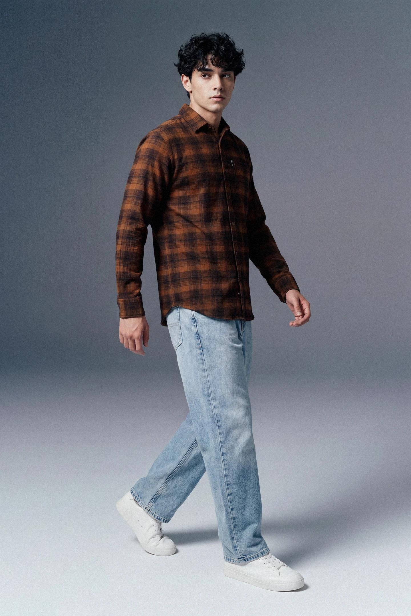 Man wearing a brown plaid shirt and light blue jeans on a gray background