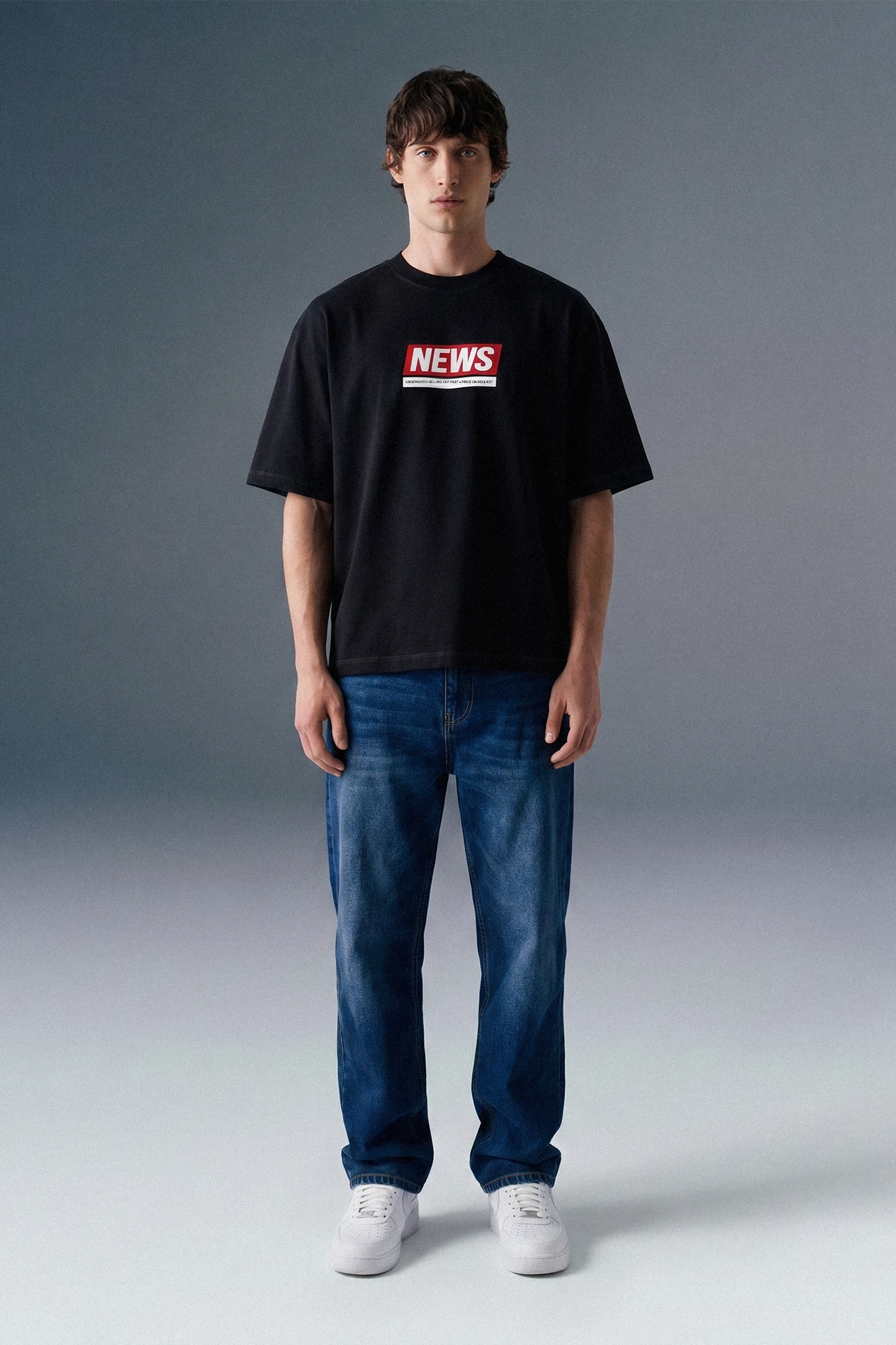 Full-length front view of a man standing straight against a grey studio backdrop, wearing a black graphic T-shirt and dark blue straight-leg jeans.