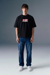 Full-length front view of a man standing straight against a grey studio backdrop, wearing a black graphic T-shirt and dark blue straight-leg jeans.