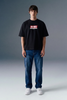 Full-length front view of a man standing straight against a grey studio backdrop, wearing a black graphic T-shirt and dark blue straight-leg jeans.