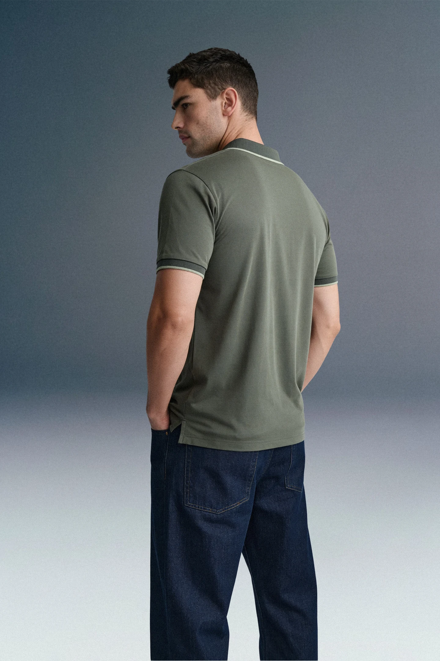 Rear view from the waist up of a man looking back over his left shoulder, wearing an olive green polo shirt with dark blue trousers.