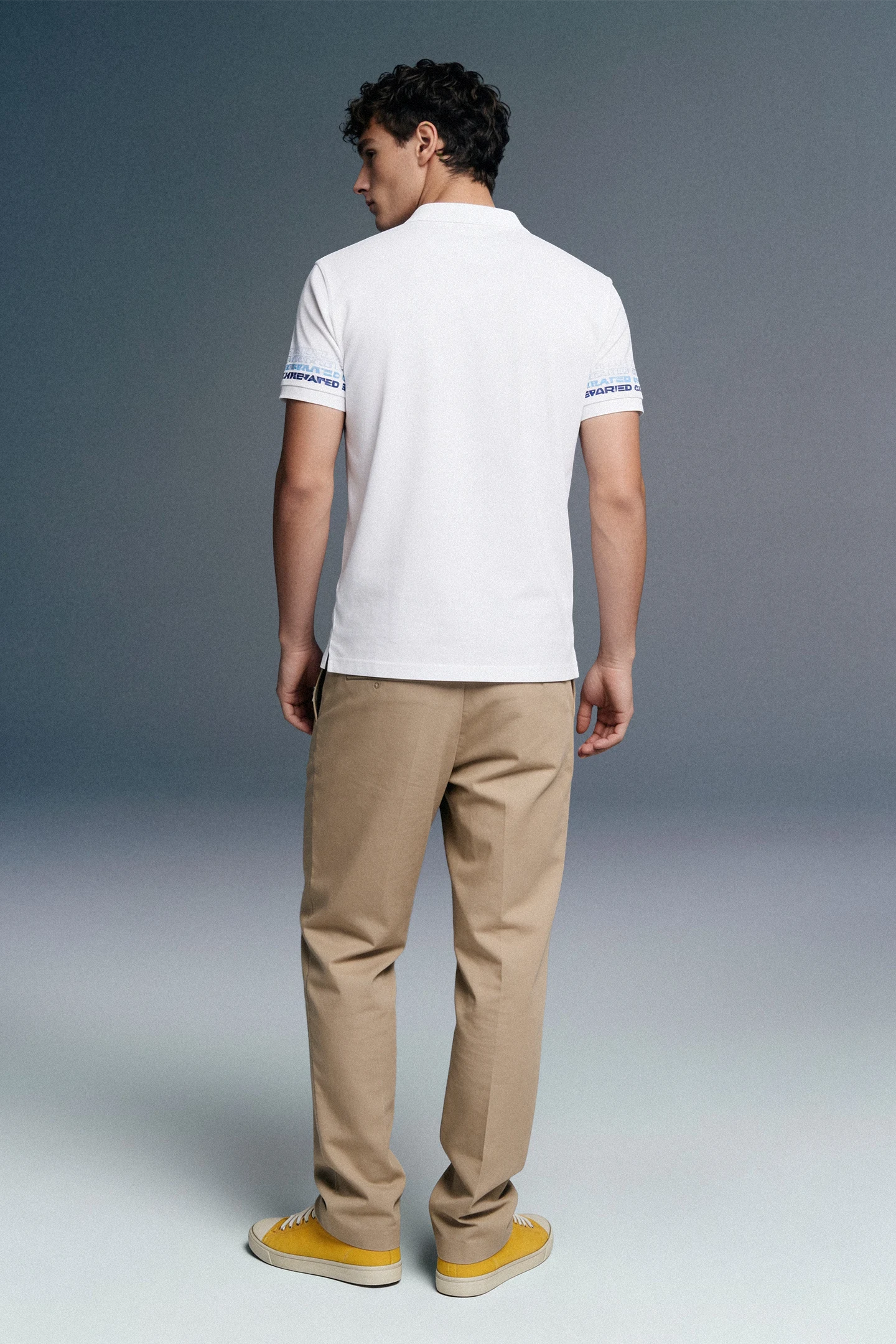 Full-length rear view of a man looking back over his shoulder, wearing a white polo shirt with blue-patterned sleeve cuffs, khaki trousers, and yellow sneakers.