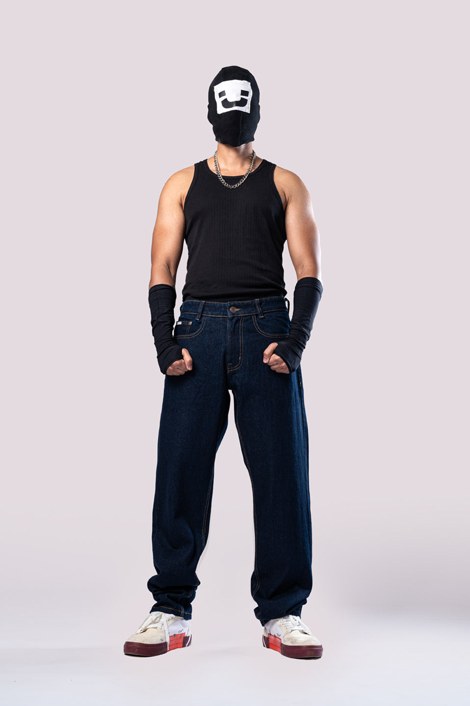 Full-body front view of a person wearing a black ski mask with a white smiley face, a black ribbed tank top, a silver chain, black arm sleeves, and dark-wash blue baggy jeans.