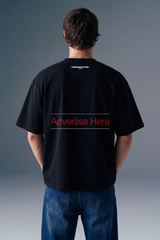 Rear view from the waist up of a man showing the back design of a black oversized T-shirt, which features subtle branding text printed across the upper back shoulders.