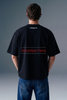 Rear view from the waist up of a man showing the back design of a black oversized T-shirt, which features subtle branding text printed across the upper back shoulders.