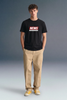 Full-length front view of a man standing facing forward, wearing a black T-shirt with a red chest graphic, khaki trousers, and yellow sneakers.