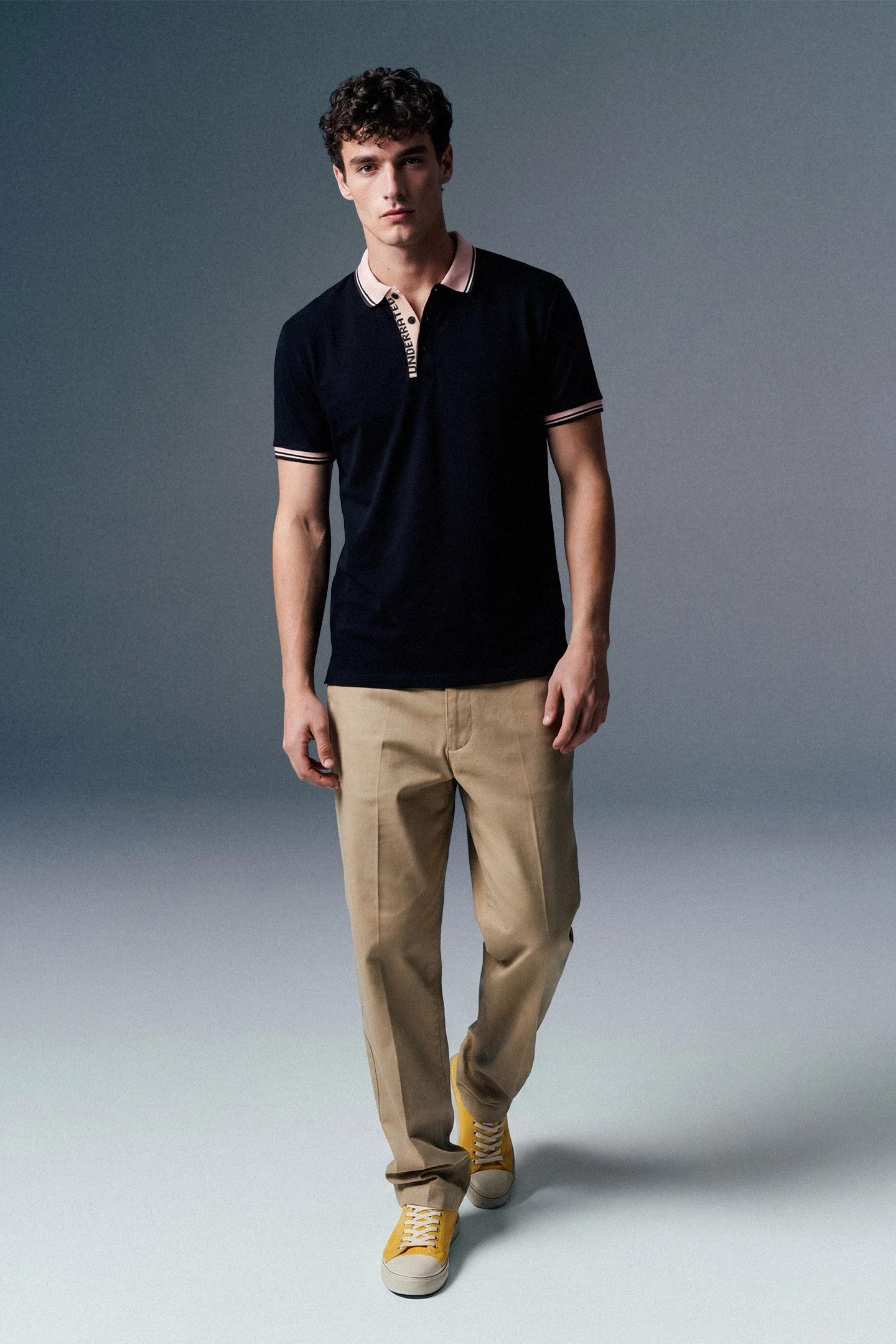 Full-length front view of a man walking slightly forward in an open-collar black polo shirt, beige pants, and yellow sneakers against a grey studio backdrop.