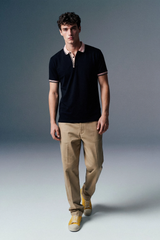 Full-length front view of a man walking slightly forward in an open-collar black polo shirt, beige pants, and yellow sneakers against a grey studio backdrop.