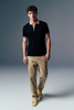 Full-length front view of a man walking slightly forward in an open-collar black polo shirt, beige pants, and yellow sneakers against a grey studio backdrop.