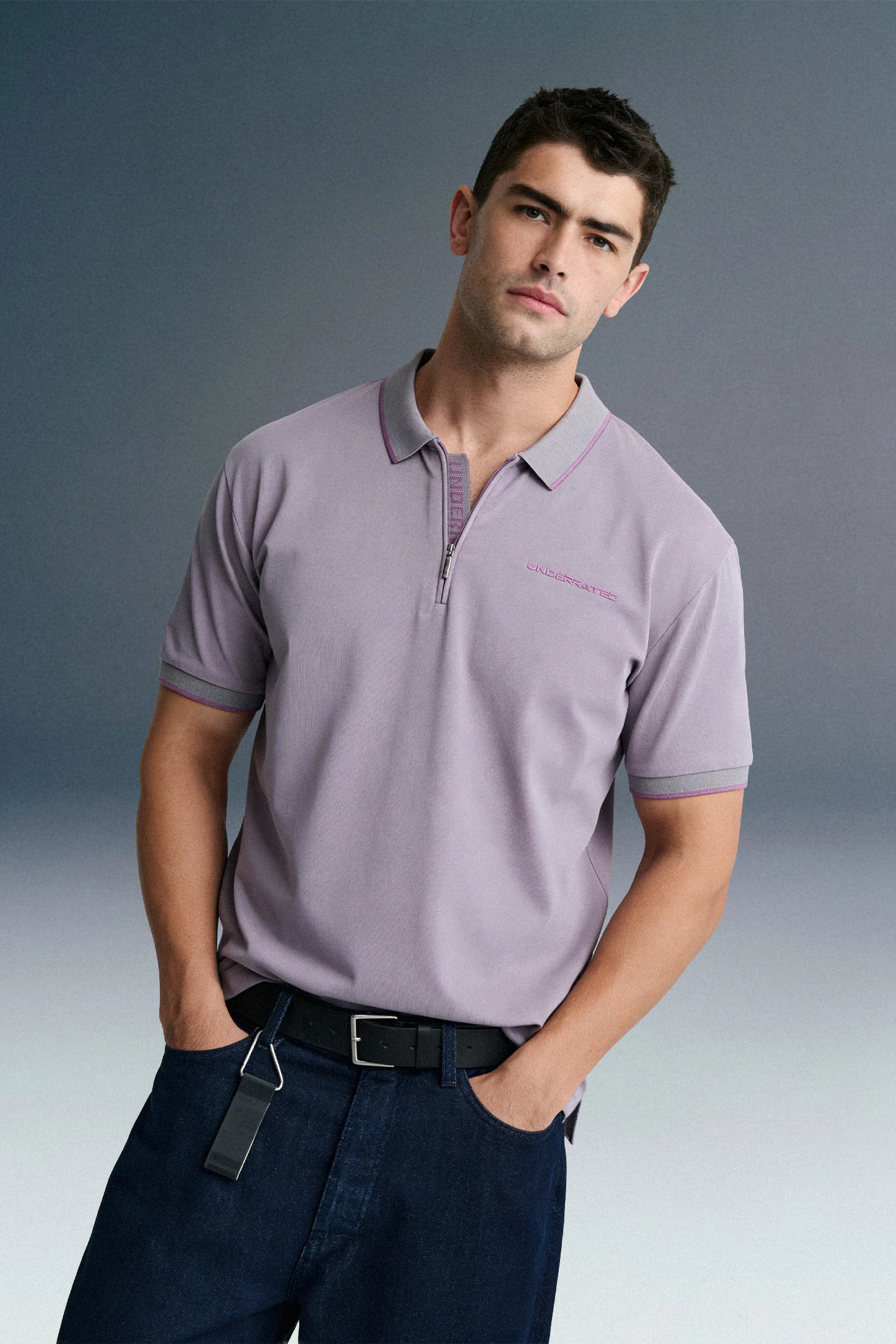 Front view from the waist up of a man leaning slightly to the side with his hands in his pockets, wearing a lavender polo shirt and dark jeans.