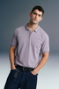Front view from the waist up of a man leaning slightly to the side with his hands in his pockets, wearing a lavender polo shirt and dark jeans.