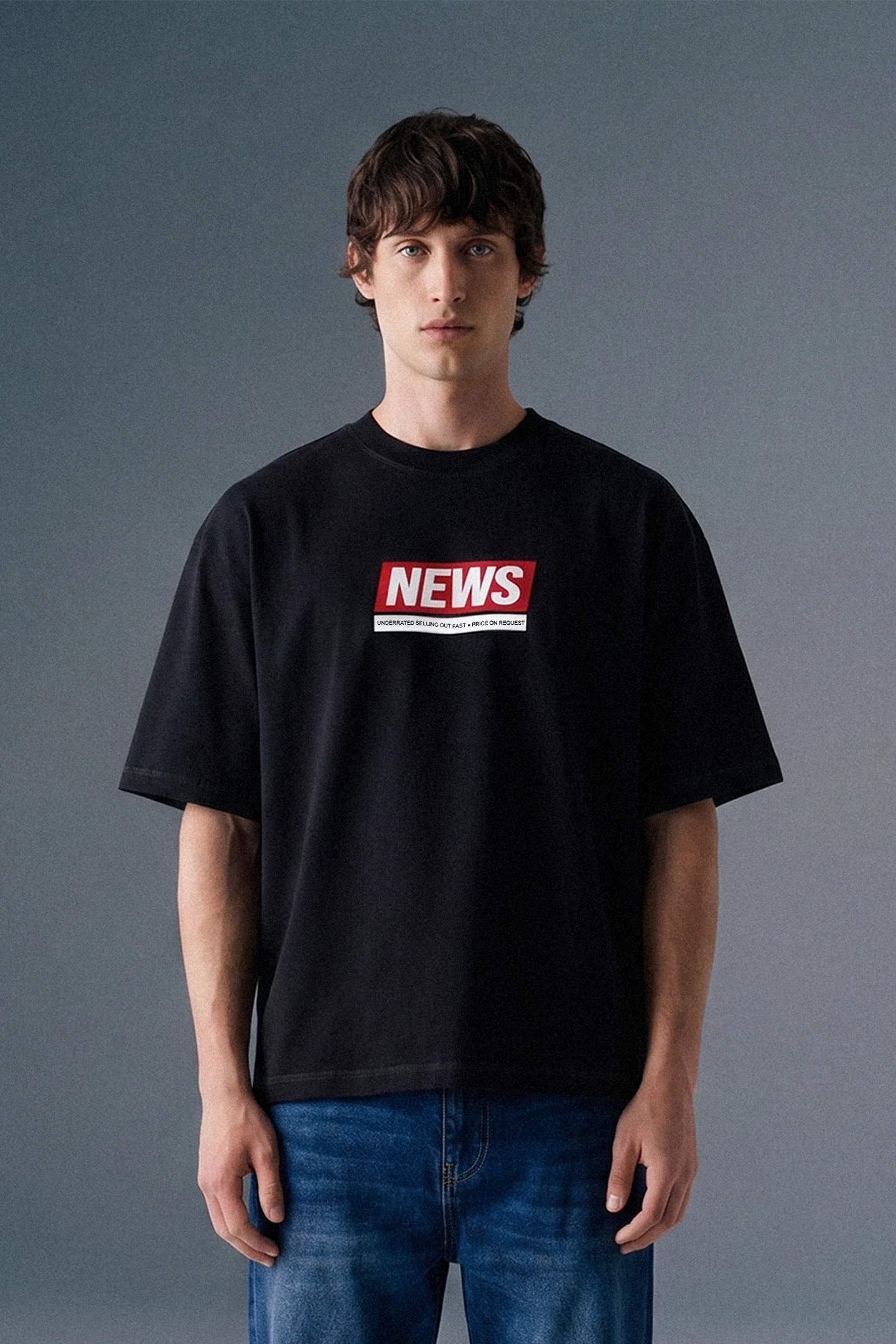 Front view from the waist up of a man looking directly forward, wearing a loose-fit black T-shirt featuring a rectangular red and white graphic on the center chest.