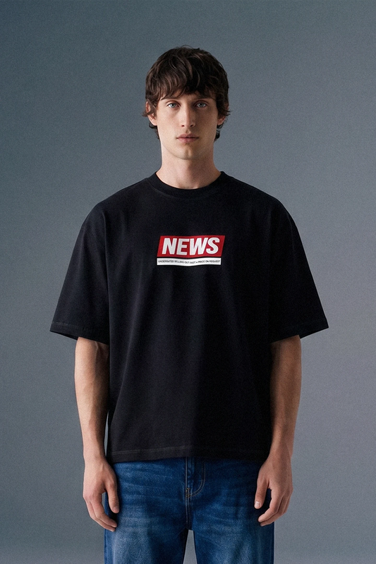 Front view from the waist up of a man looking directly forward, wearing a loose-fit black T-shirt featuring a rectangular red and white graphic on the center chest.