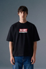 Front view from the waist up of a man looking directly forward, wearing a loose-fit black T-shirt featuring a rectangular red and white graphic on the center chest.
