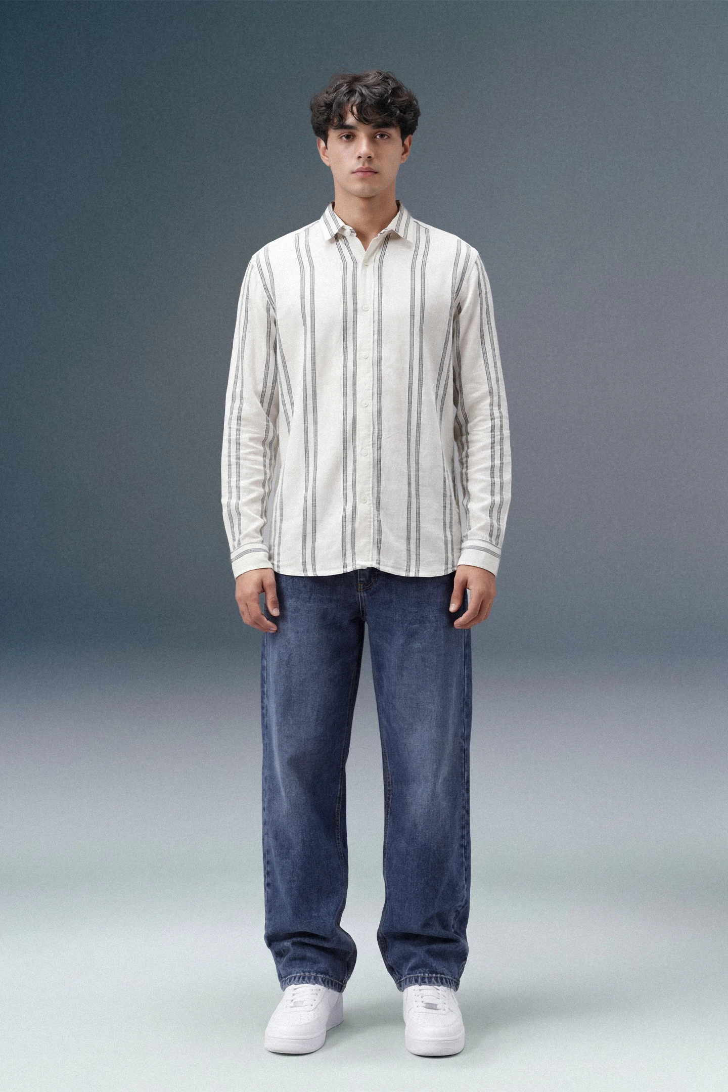 Man wearing a striped shirt and jeans standing against a gray background
