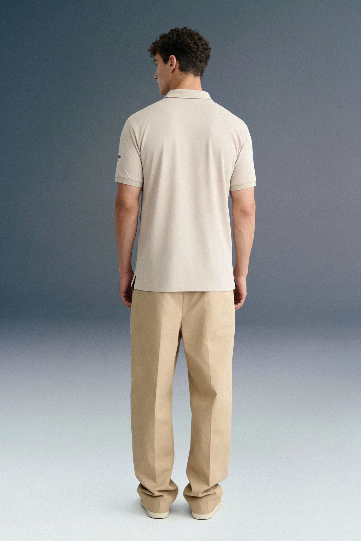Rear view from the waist up of a man standing still against a grey studio background, highlighting the back structure and shoulders of a dark navy blue polo shirt.