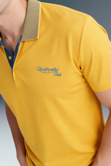 Side profile close-up focusing on the shoulder and collar of a mustard yellow polo shirt, showing a khaki knit collar texture and blue graphic branding on the left chest.