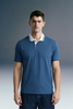 A three-quarter front shot of a man standing still, wearing a slate blue polo shirt with white trim on the collar and cuffs, paired with dark trousers.