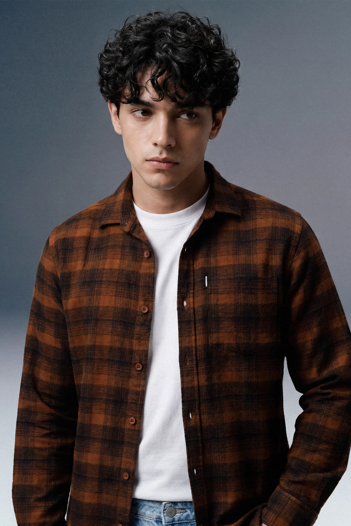 Man wearing a brown plaid shirt over a white t-shirt against a gray background