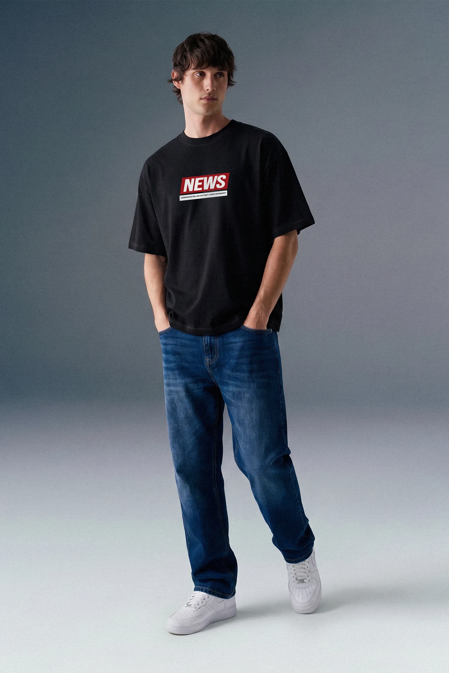 Full-length front view of a man walking slightly forward with his hands in his pockets, wearing an oversized black T-shirt with a red and white chest graphic, blue jeans, and light-colored sneakers.