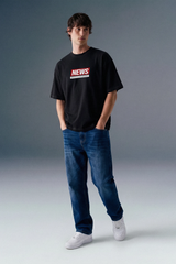 Full-length front view of a man walking slightly forward with his hands in his pockets, wearing an oversized black T-shirt with a red and white chest graphic, blue jeans, and light-colored sneakers.